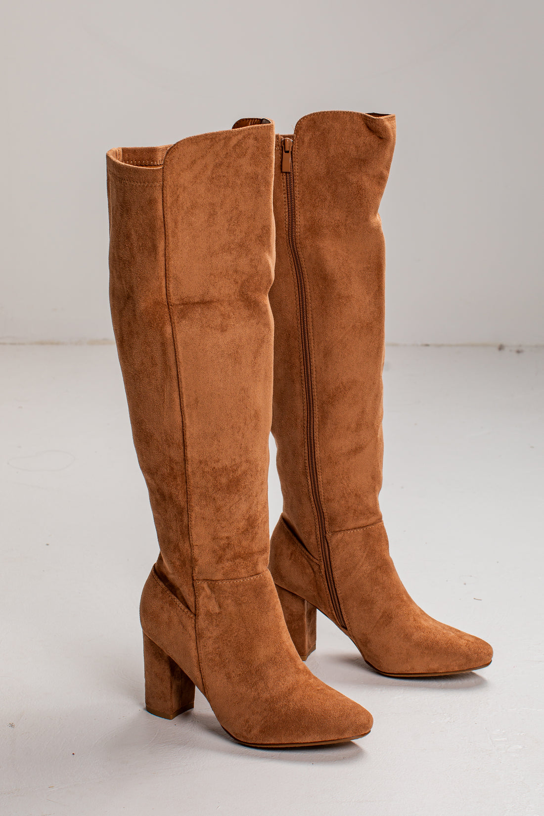 A pair of knee-high brown suede boots with a block heel and pointed toe, shown on a light gray studio background.