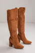 A pair of knee-high brown suede boots with a block heel and pointed toe, shown on a light gray studio background.