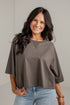 A woman with long dark hair smiling while wearing a loose-fitting, short-sleeve taupe-brown top and light-wash blue jeans against a light gray wall.