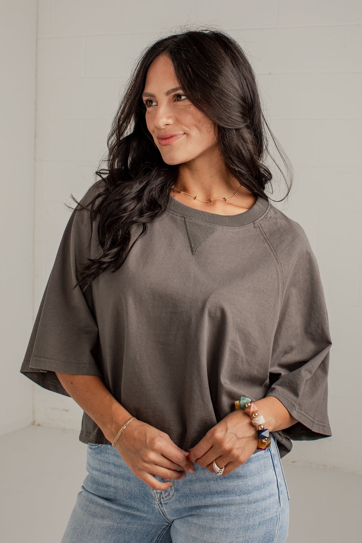A woman wearing a loose-fitting, short-sleeve crewneck top in a muted taupe-brown color, paired with light-wash blue jeans, standing against a light gray wall.