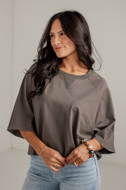 A woman wearing a loose-fitting, short-sleeve crewneck top in a muted taupe-brown color, paired with light-wash blue jeans, standing against a light gray wall.