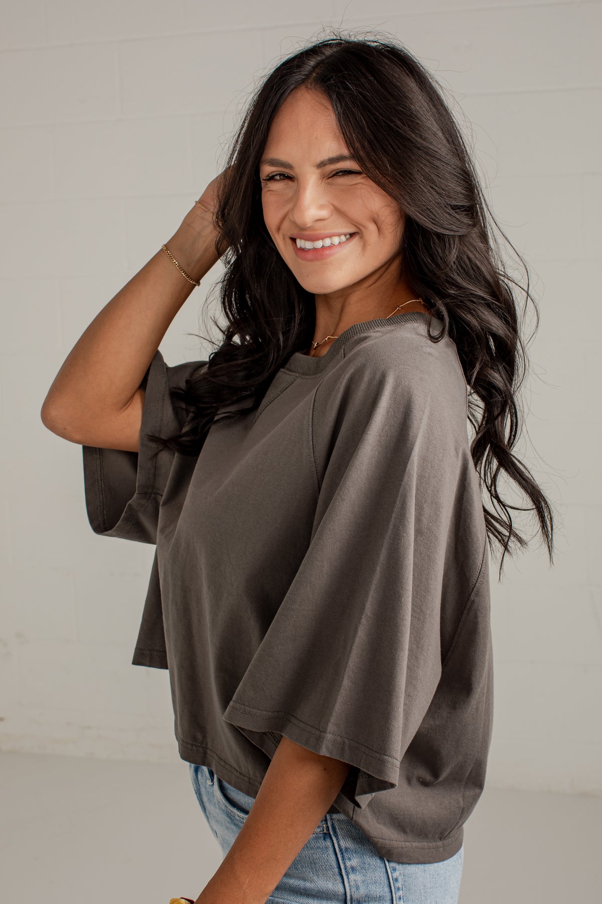 A woman with long dark hair smiling and posing in a loose-fitting, oversized charcoal gray t-shirt with wide three-quarter sleeves, paired with light blue jeans against a white wall background.