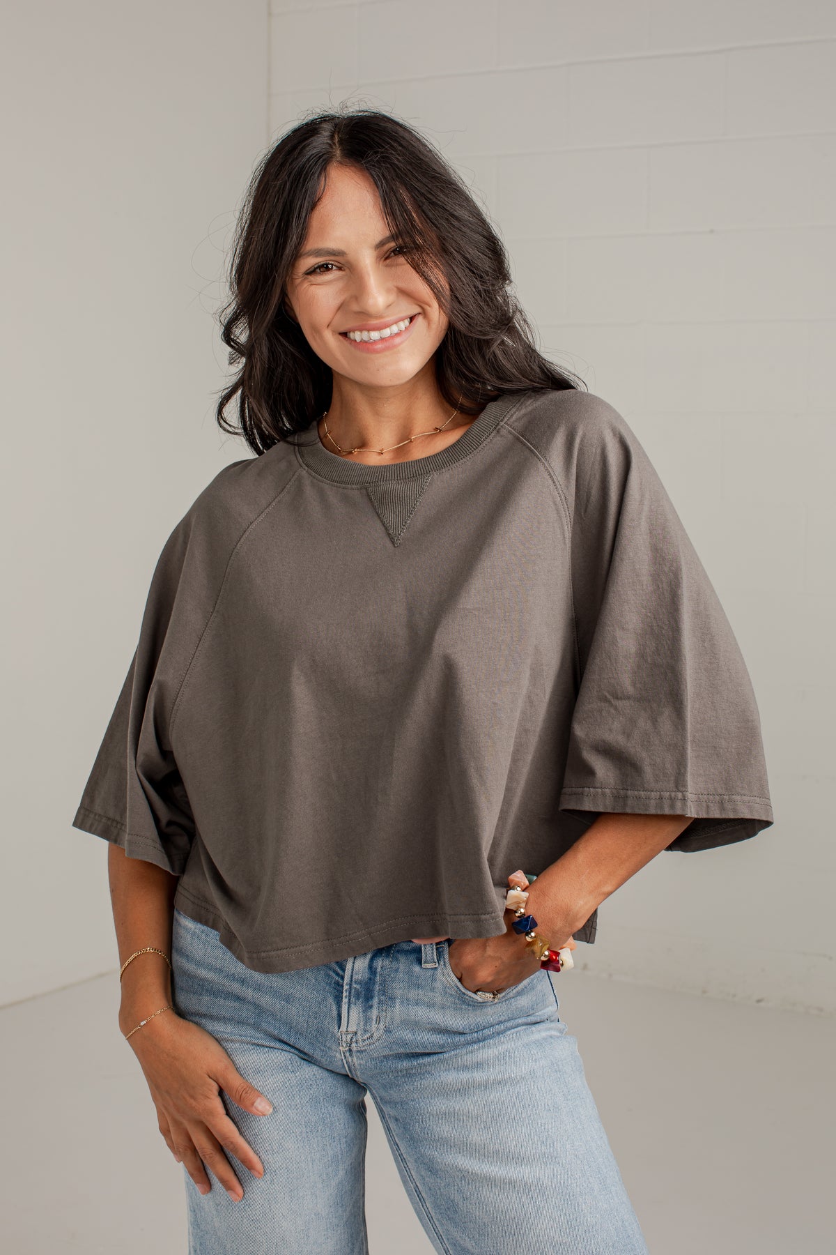A woman wearing a loose-fitting, short-sleeve taupe-brown top and light-wash blue jeans, standing against a light gray wall.