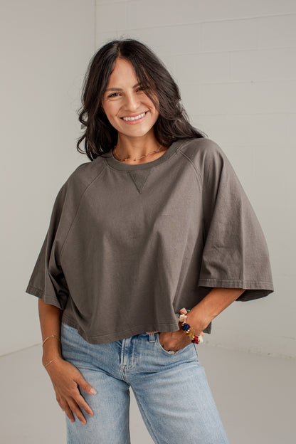 A woman wearing a loose-fitting, short-sleeve taupe-brown top and light-wash blue jeans, standing against a light gray wall.