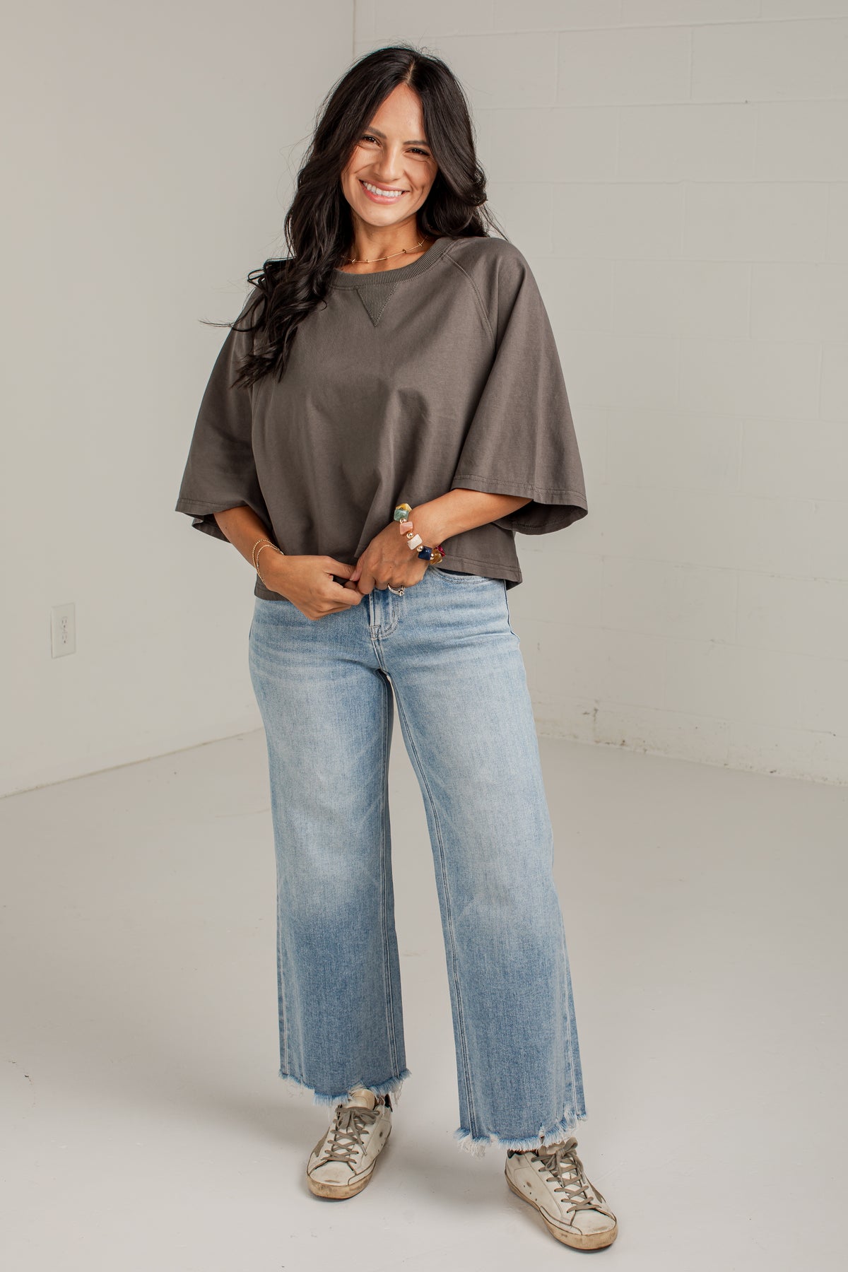 A woman modeling a loose, short-sleeve taupe-brown top with wide bell sleeves, paired with light-wash wide-leg jeans and beige sneakers, standing against a white studio wall.