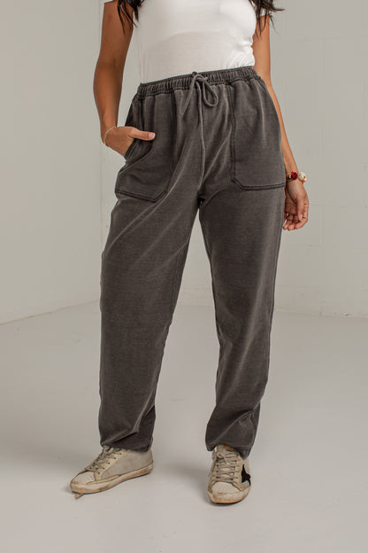 A person wearing dark gray drawstring sweatpants with side pockets, paired with a white sleeveless top and beige sneakers, standing against a plain light gray studio background.
