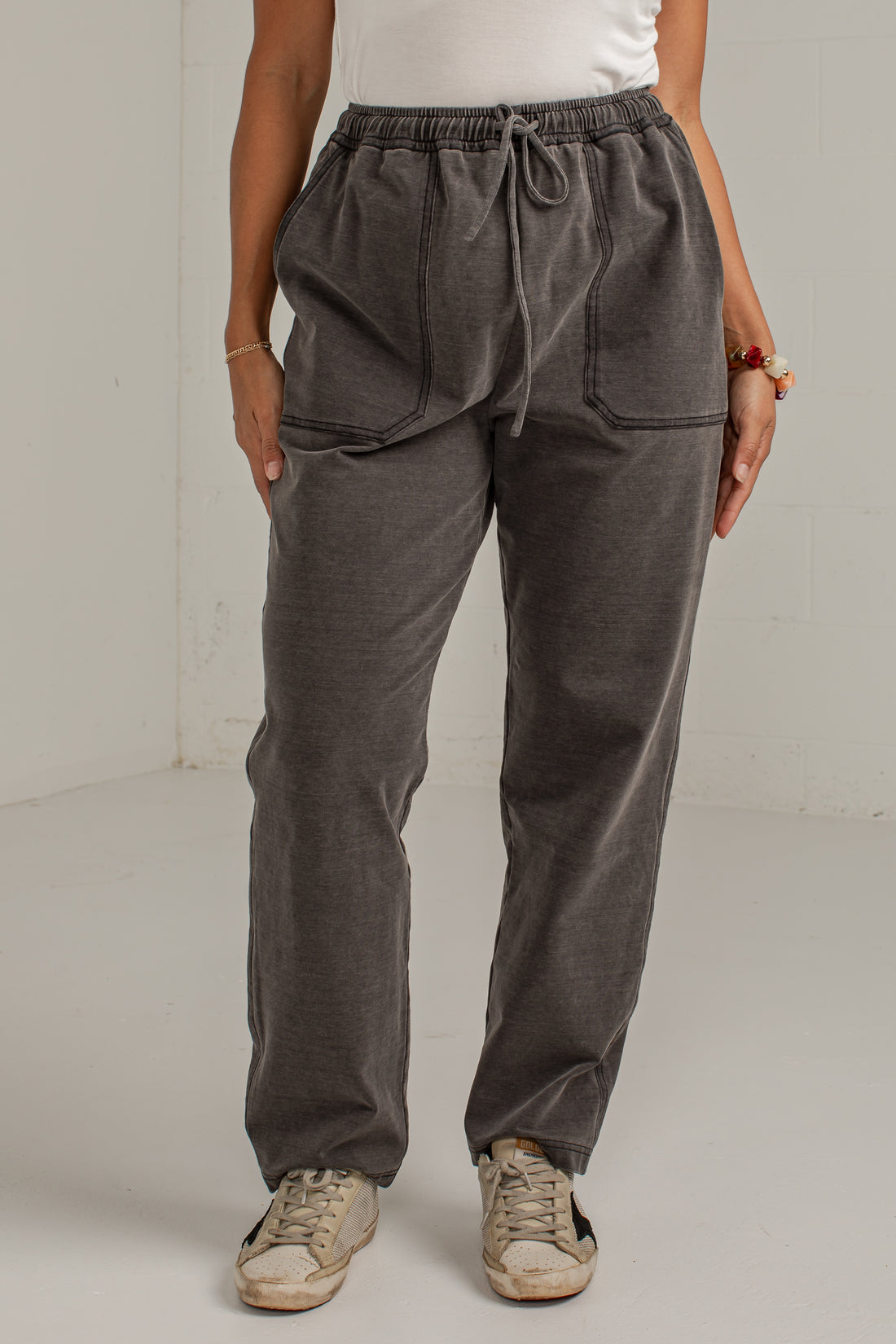 A person wearing dark gray drawstring sweatpants with large front patch pockets, paired with a white top and beige sneakers, standing against a plain light gray wall.