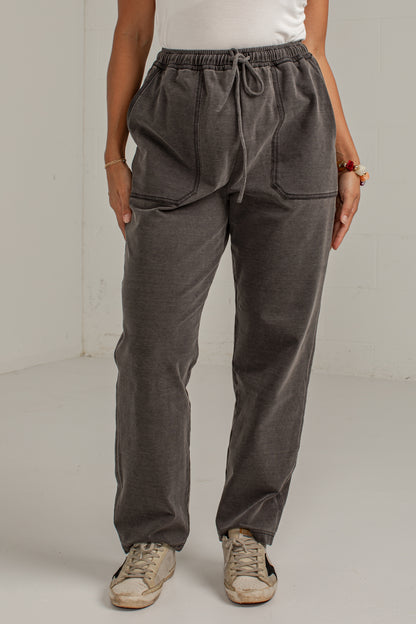 A person wearing dark gray drawstring sweatpants with large front patch pockets, paired with a white top and beige sneakers, standing against a plain light gray wall.
