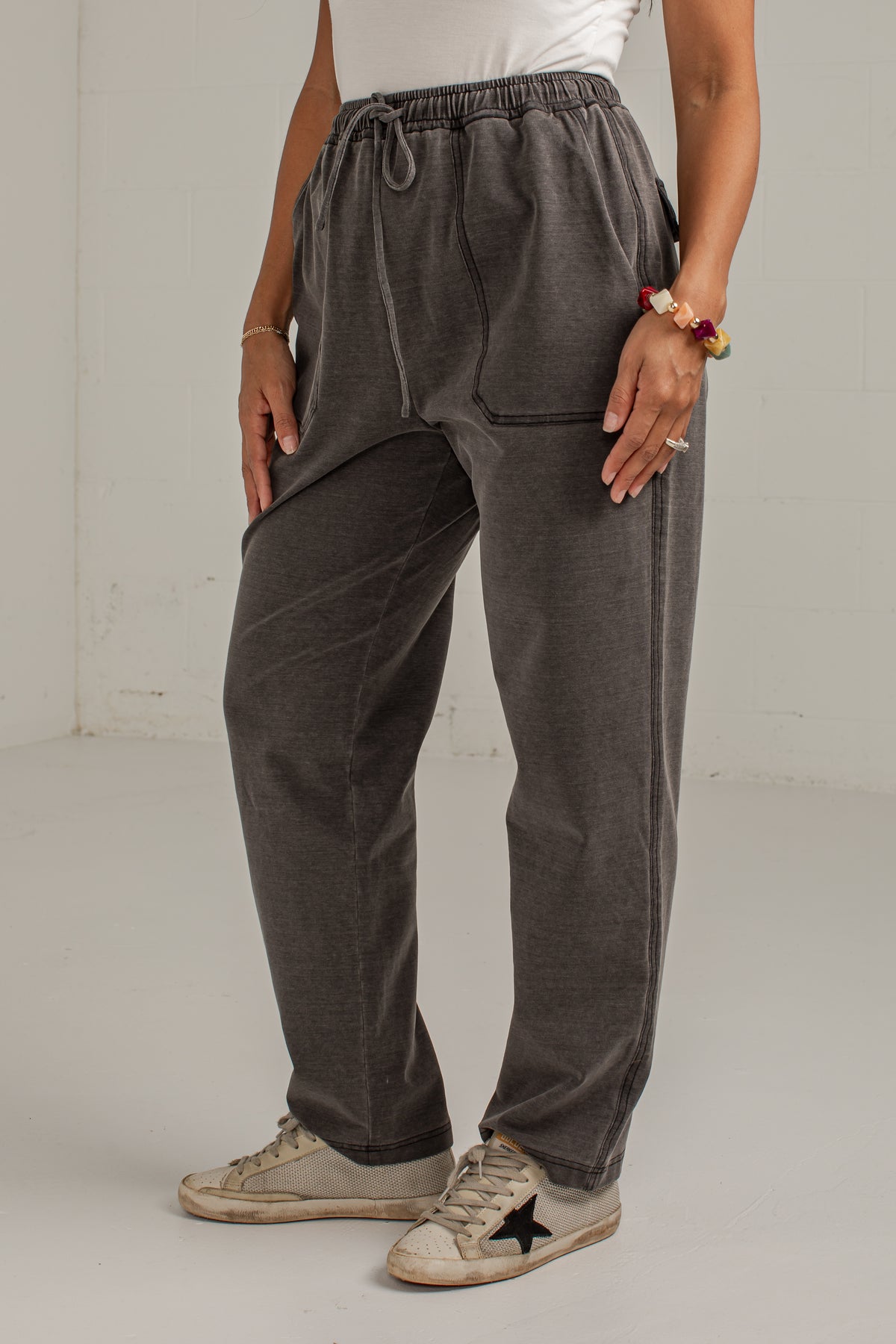 A person wearing dark gray drawstring sweatpants with side pockets, paired with a white top and beige sneakers, standing against a plain light gray wall.
