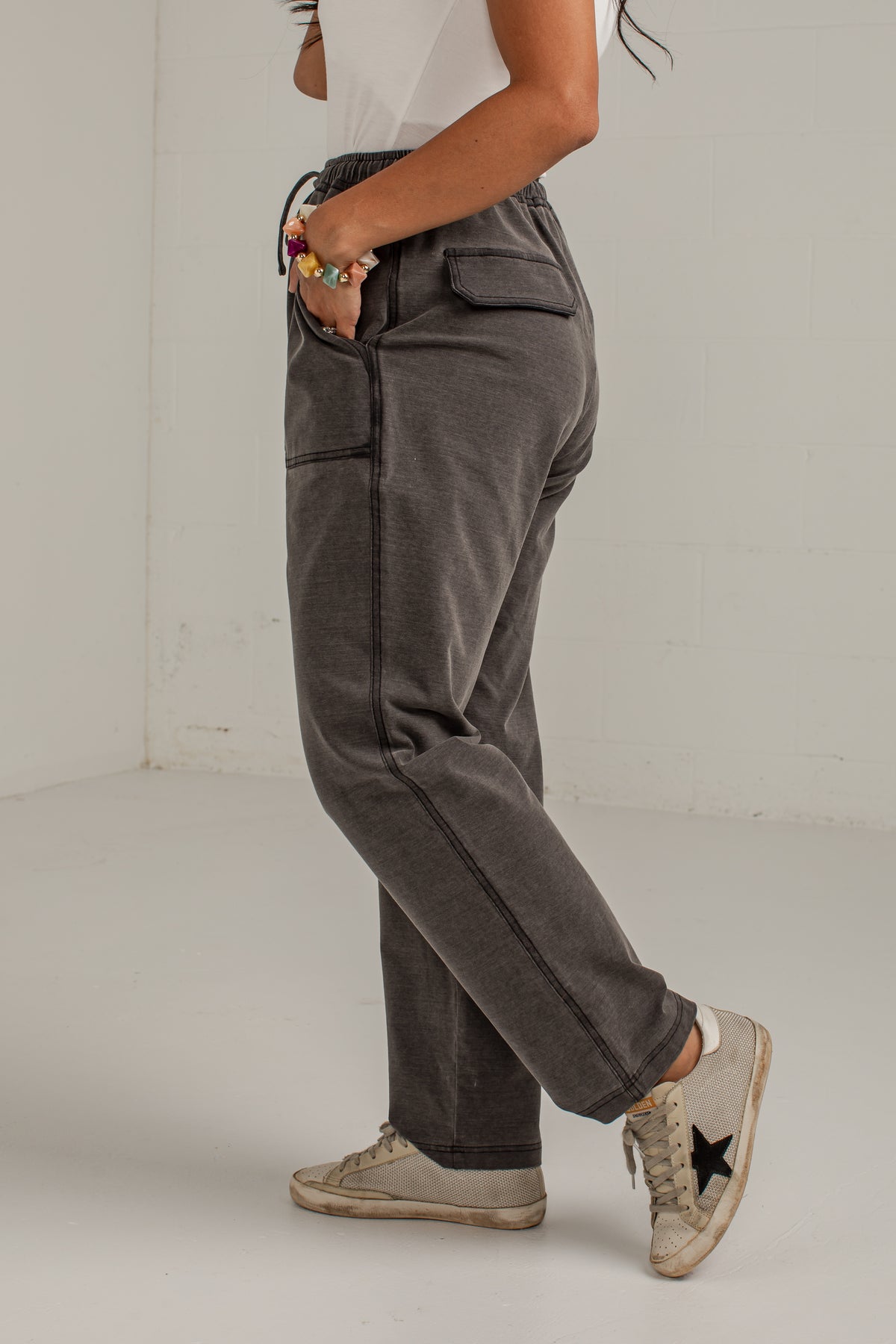 A person wearing dark gray washed corduroy pants with side pockets and a drawstring waist, paired with a white top and beige sneakers, standing in a minimalist white studio.