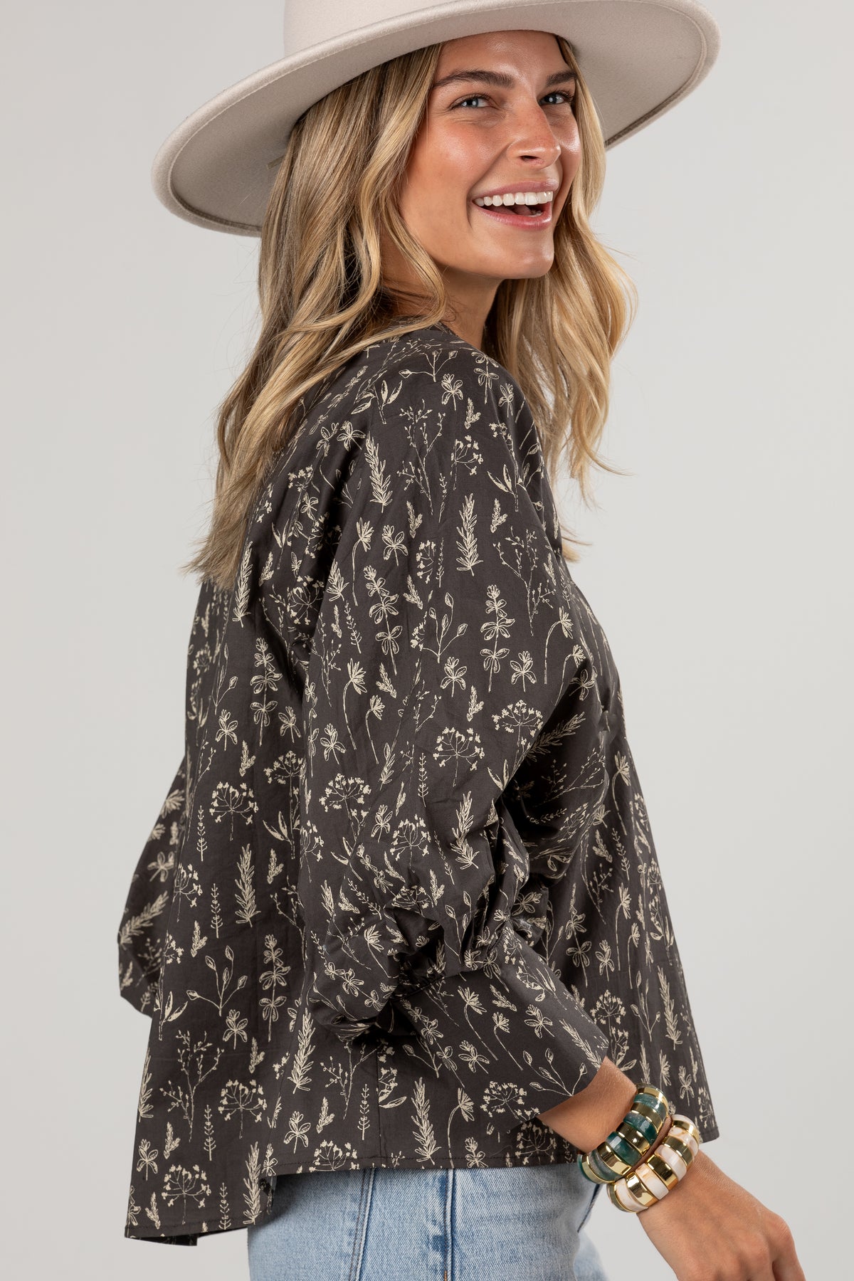 A woman wearing a dark brown long-sleeve blouse with a beige botanical floral pattern, styled with a wide-brimmed cream hat and light blue jeans against a light gray studio background.