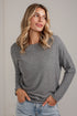 A woman wearing a heather gray long-sleeve knit sweater and light blue jeans, standing against a light off-white paneled wall.