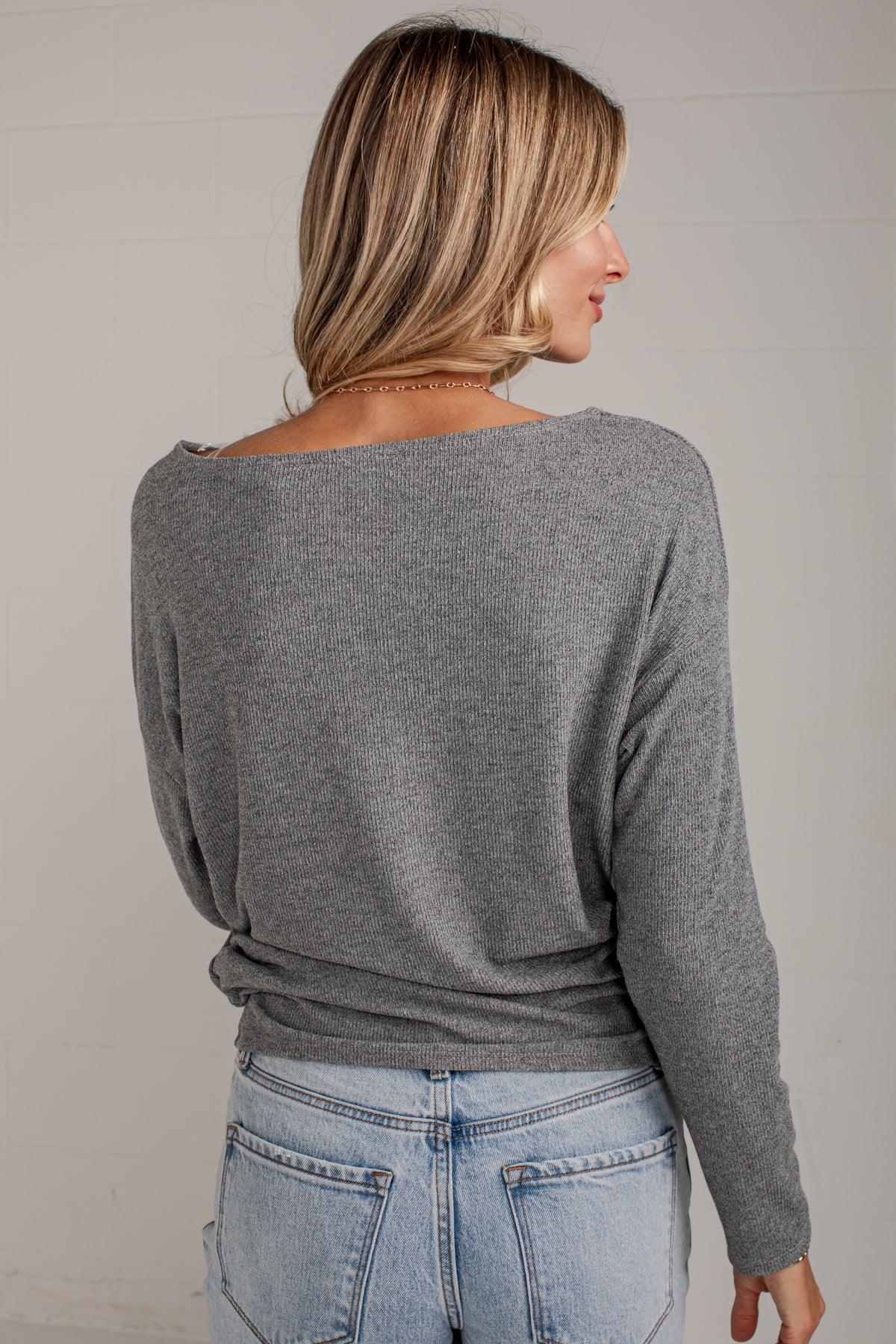 A woman shown from the back wearing a heather gray long-sleeve knit top with a wide boat neckline, paired with light blue jeans, against a light gray wall.