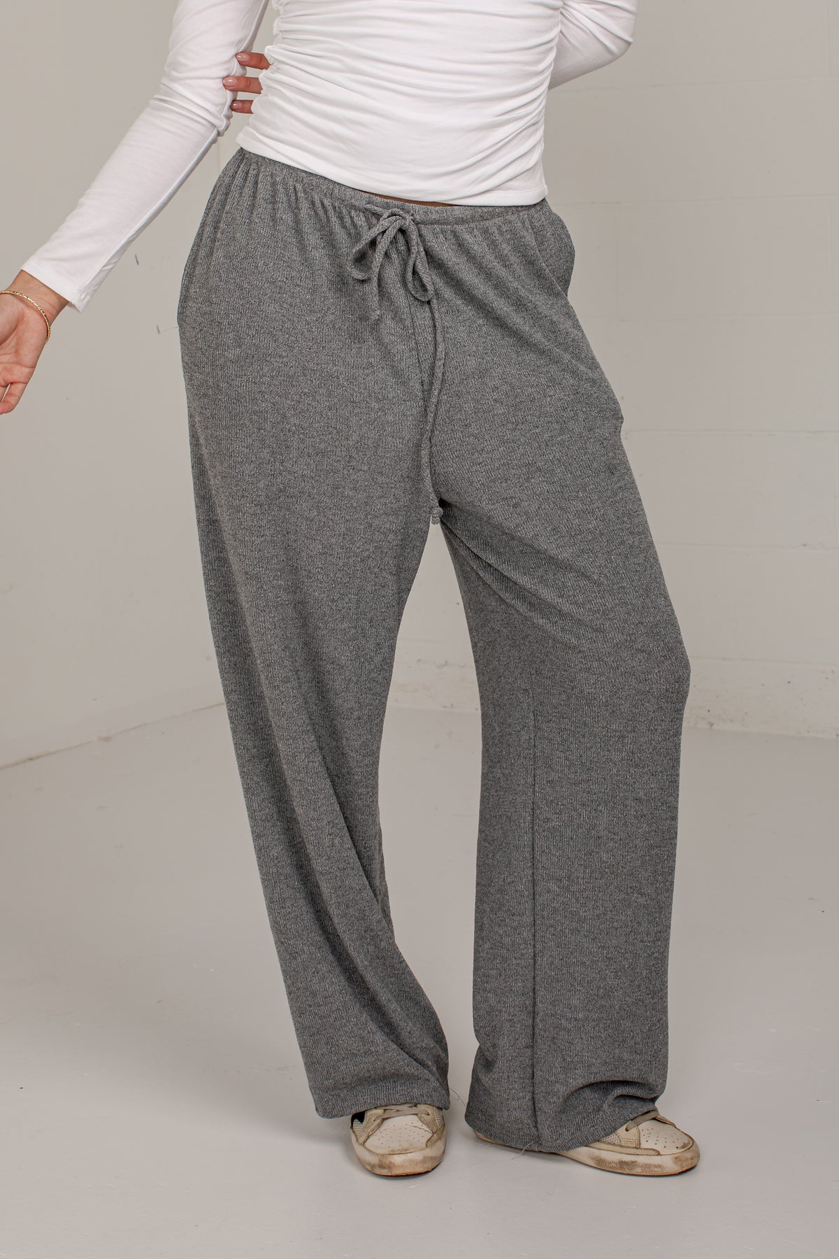 A person wearing loose, wide-leg gray drawstring sweatpants with a white long-sleeve top and beige shoes, photographed in a plain indoor studio setting.