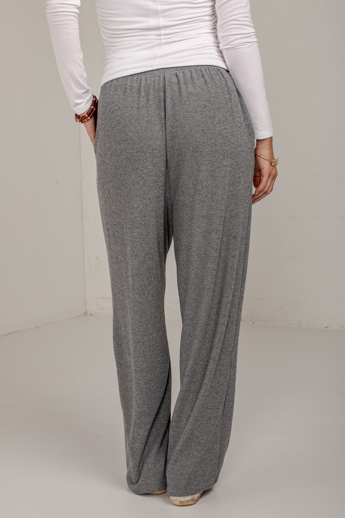 A person shown from the back wearing loose, wide-leg gray knit pants and a white long-sleeve top in a plain indoor setting.