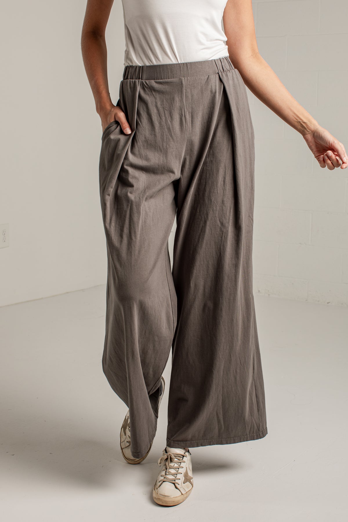 A person wearing wide-leg pleated trousers in a taupe-gray color, styled with a white sleeveless top and beige sneakers, standing against a light gray wall.