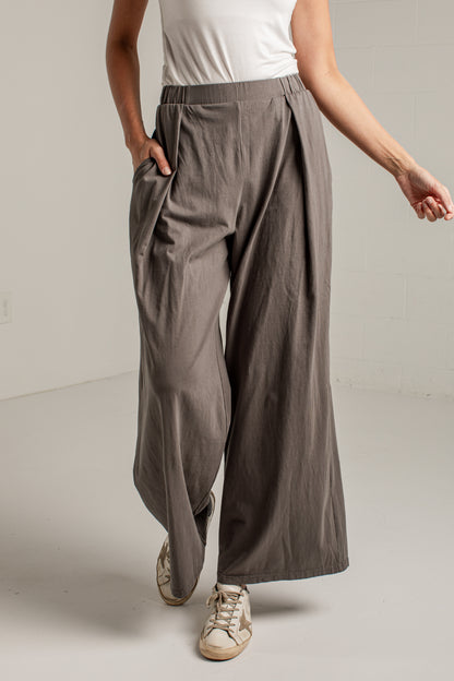 A person wearing wide-leg pleated trousers in a taupe-gray color, styled with a white sleeveless top and beige sneakers, standing against a light gray wall.