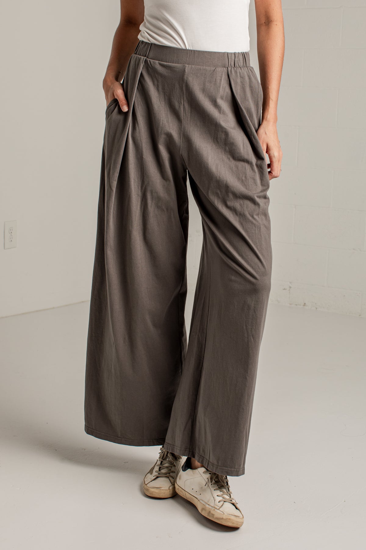 A person wearing loose, wide-leg pleated pants in a muted taupe-gray color, paired with a white top and beige sneakers, standing in a minimalist studio setting.