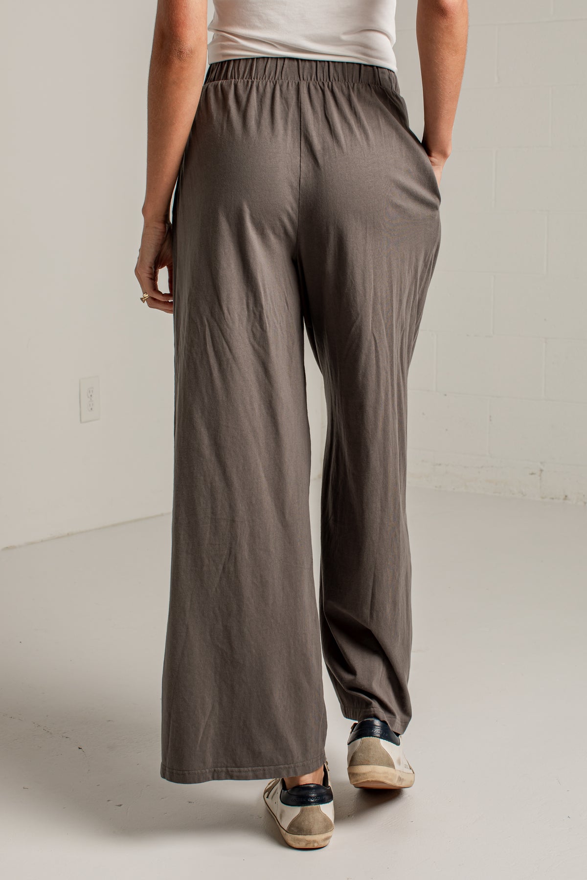 A person shown from the back wearing loose, wide-leg taupe pants, a white top, and beige sneakers in a bright studio setting.