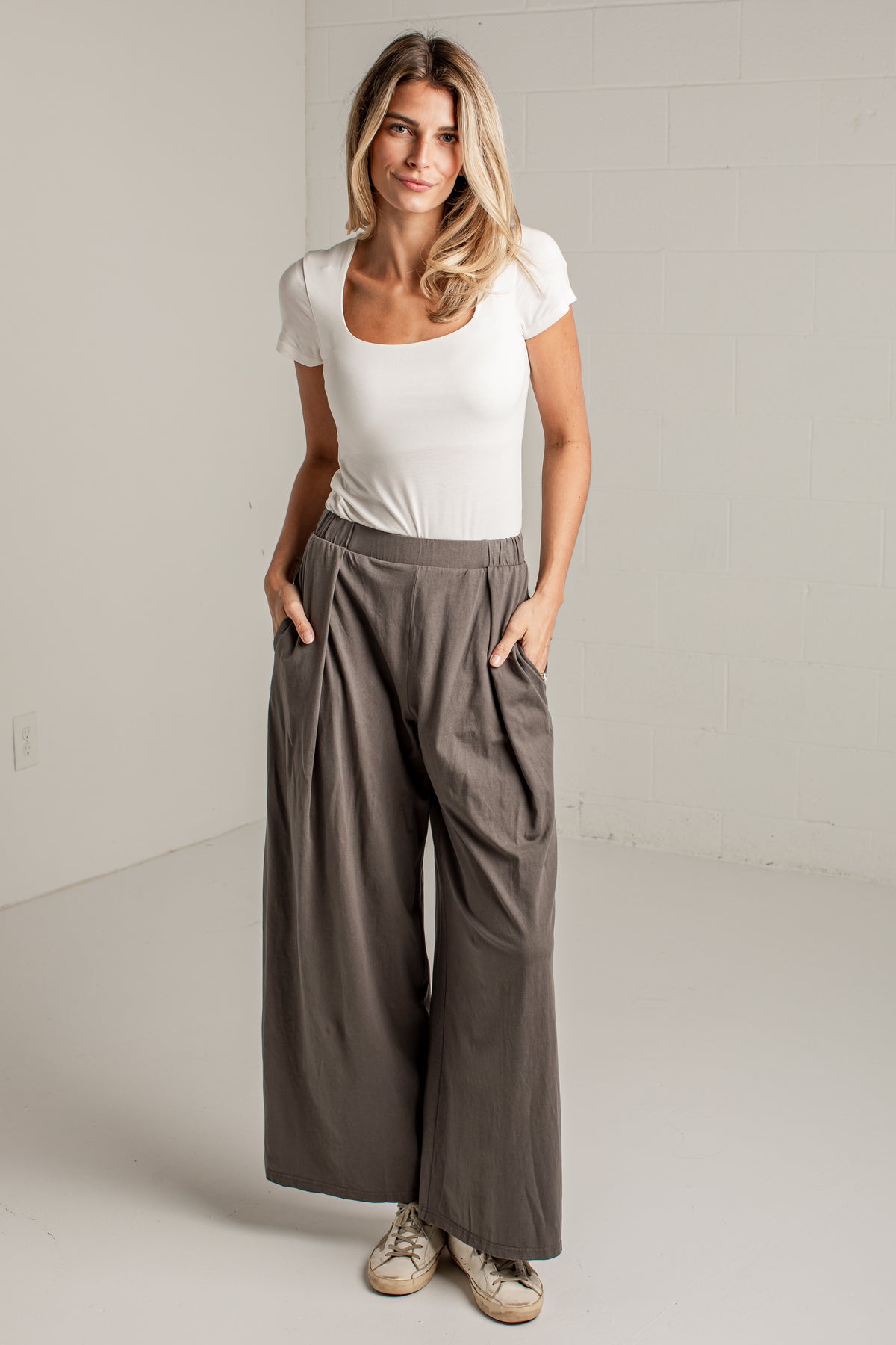 A woman modeling a pair of wide-leg taupe pants with a white scoop-neck t-shirt, standing in a bright, neutral studio setting.