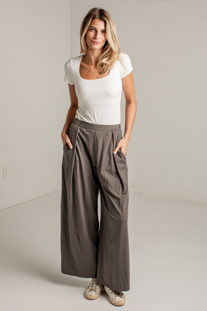 A woman modeling a pair of wide-leg taupe pants with a white scoop-neck t-shirt, standing in a bright, neutral studio setting.