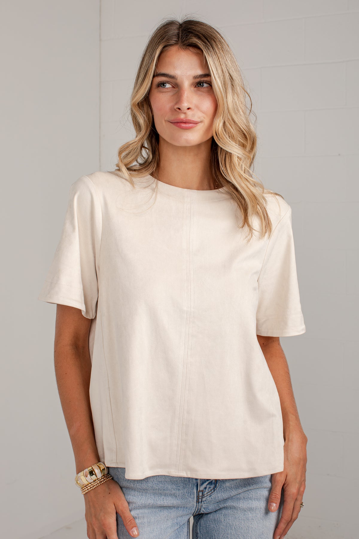 A woman wearing a cream-colored short-sleeve top and light blue jeans, standing against a light gray wall.