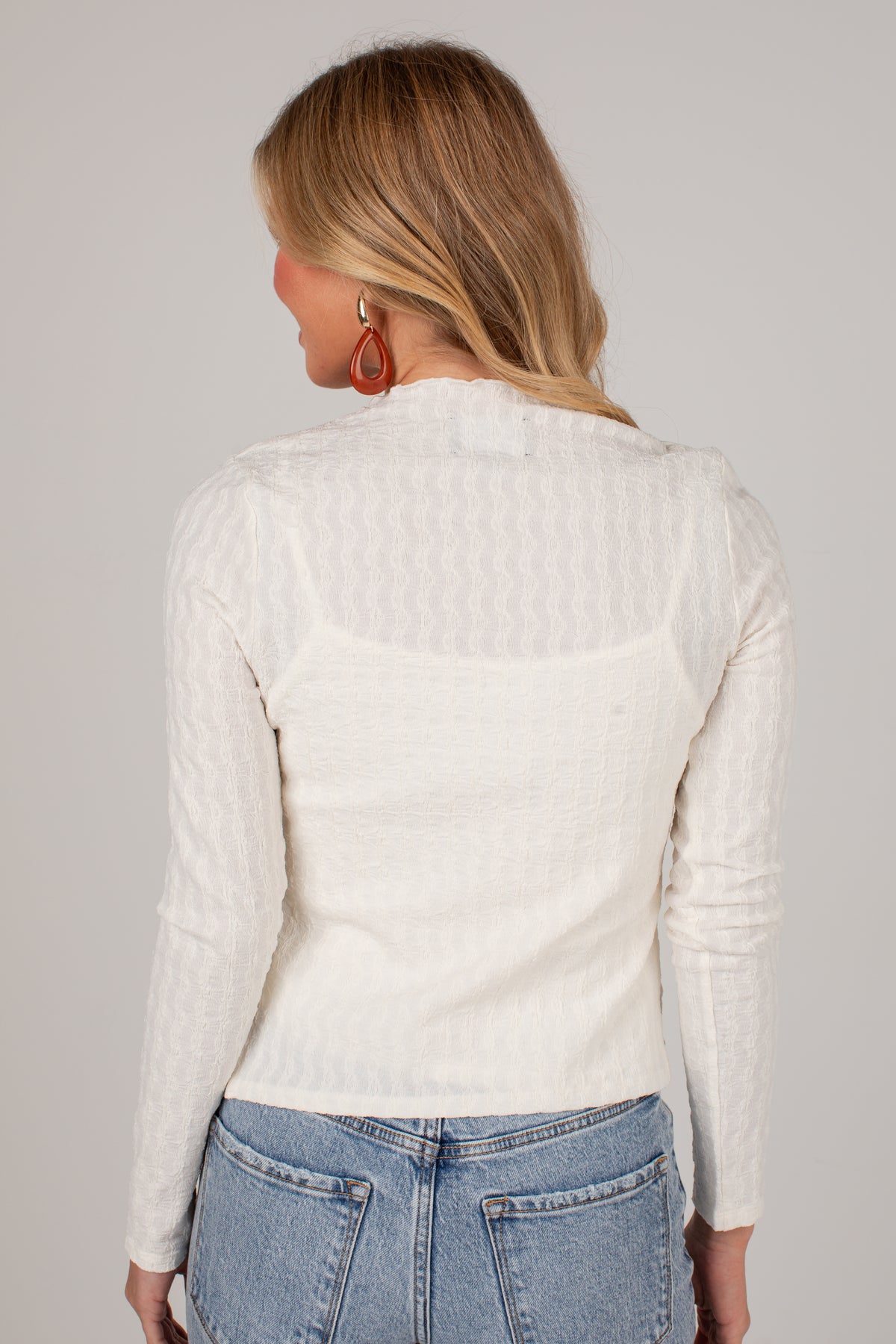 A woman shown from the back wearing a cream-colored textured knit long-sleeve top and light blue jeans against a solid light gray background.