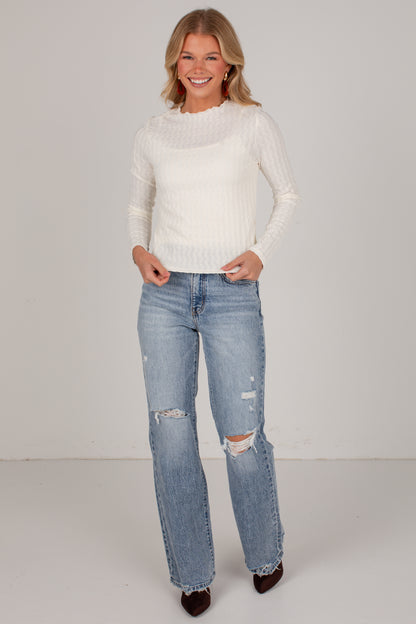 A woman modeling a white ribbed knit mock-neck sweater and light-wash blue distressed jeans with ripped knees, standing against a plain light gray studio background.