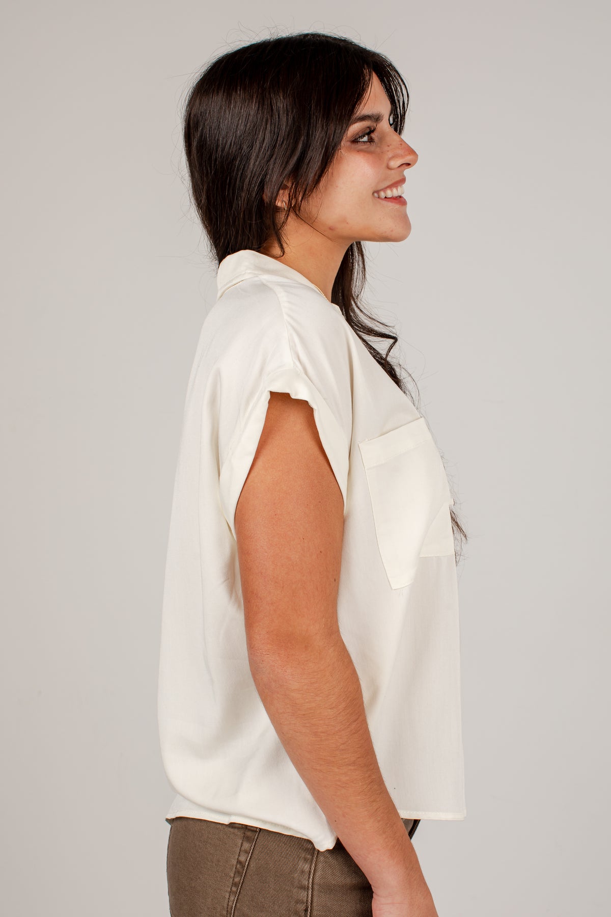 A woman shown in side profile wearing a short-sleeve white blouse with a chest pocket, paired with brown pants, against a plain light gray studio background.