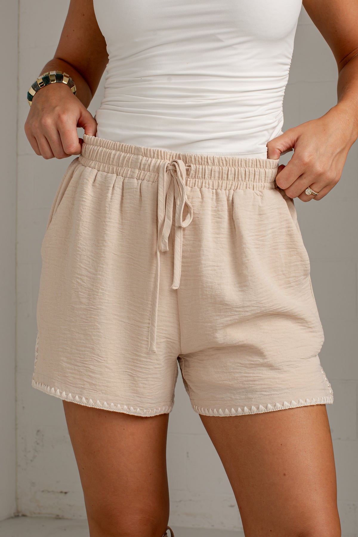 Beige textured drawstring shorts worn by a model, paired with a white tank top, shown from the chest down against a light gray wall.