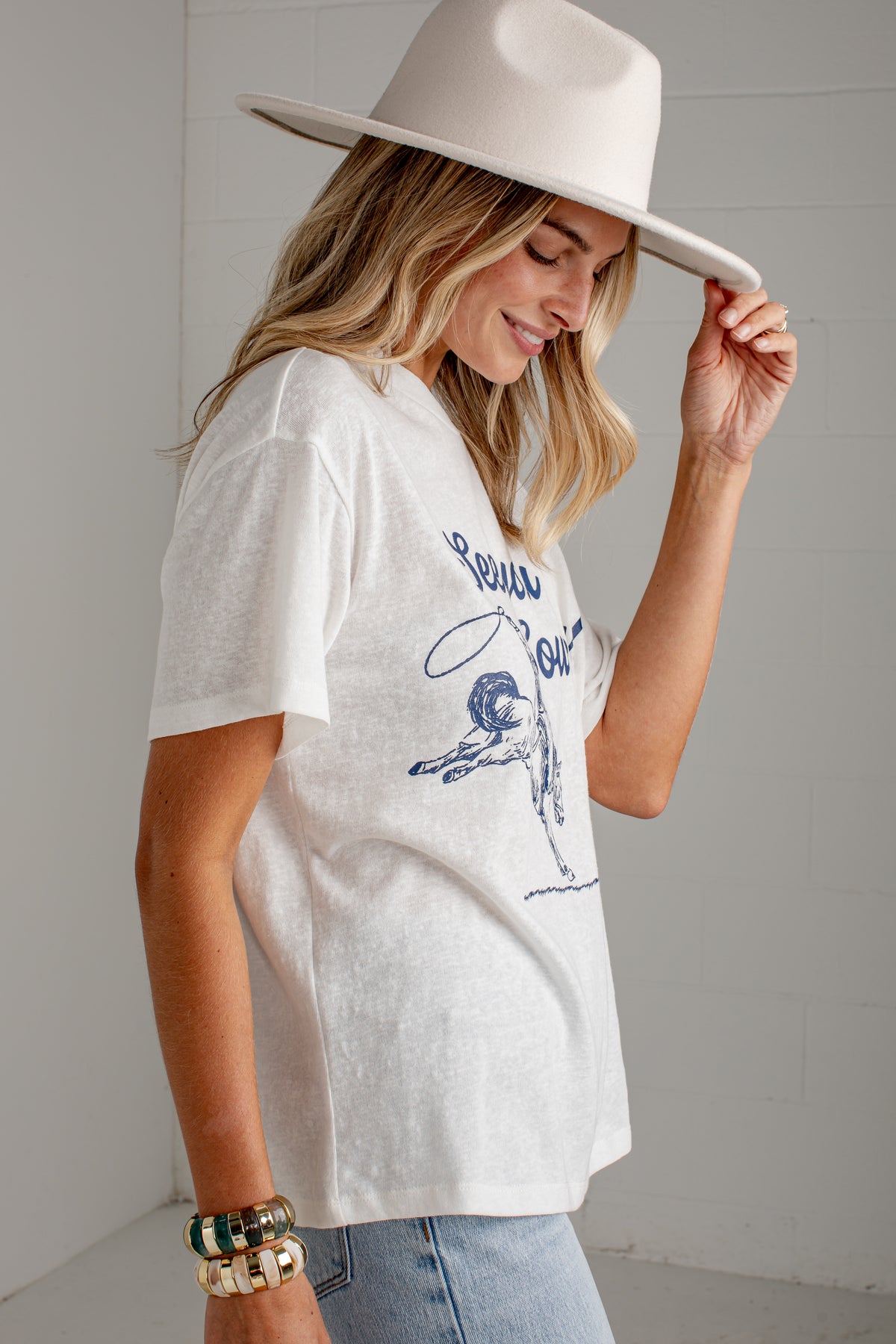 A woman wearing a white graphic t-shirt with a blue vintage-style print, paired with light-wash jeans and a wide-brimmed cream hat, posing in a bright, modern room.