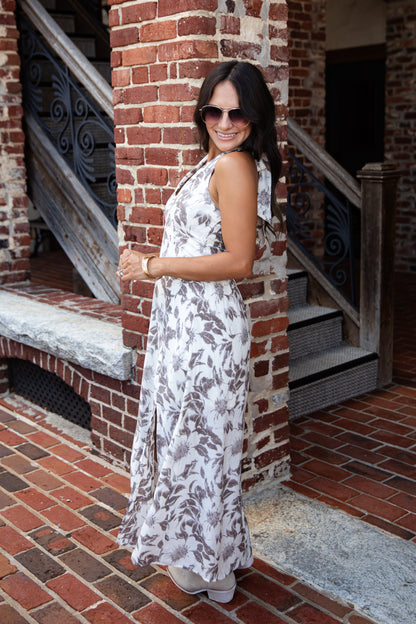 A woman wearing a sleeveless white maxi dress with a brown floral pattern, standing on a brick patio in front of a brick building with wooden railings.