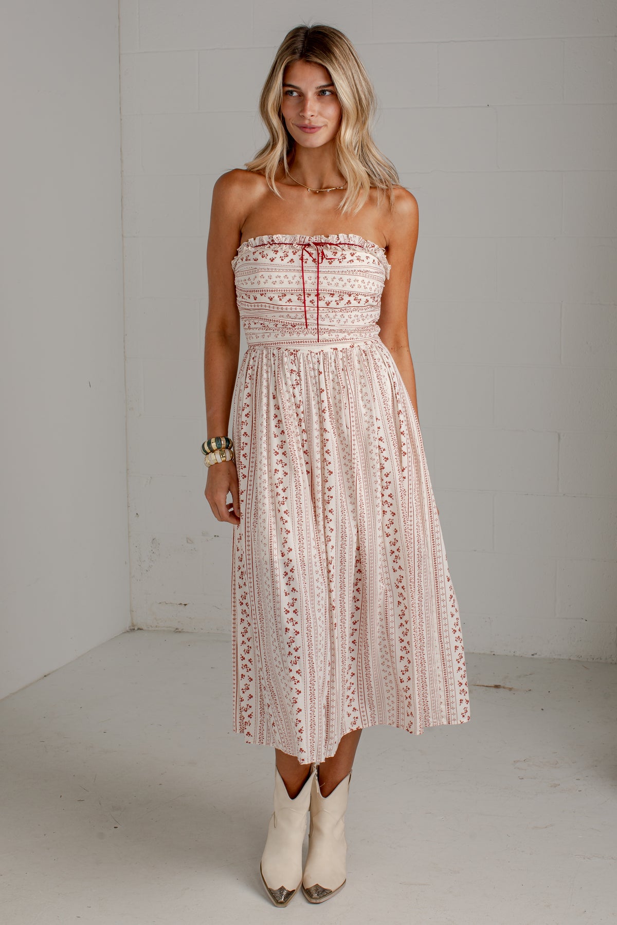 A woman modeling a strapless midi dress with a cream and pink floral pattern, featuring a tie-neck detail and a flared skirt, paired with white cowboy boots against a white brick wall background.