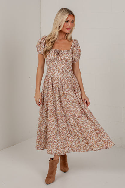 A woman modeling a midi-length puff-sleeve dress with a small floral print in beige, brown, and muted pink tones, paired with tan ankle boots, set against a plain off-white studio background.