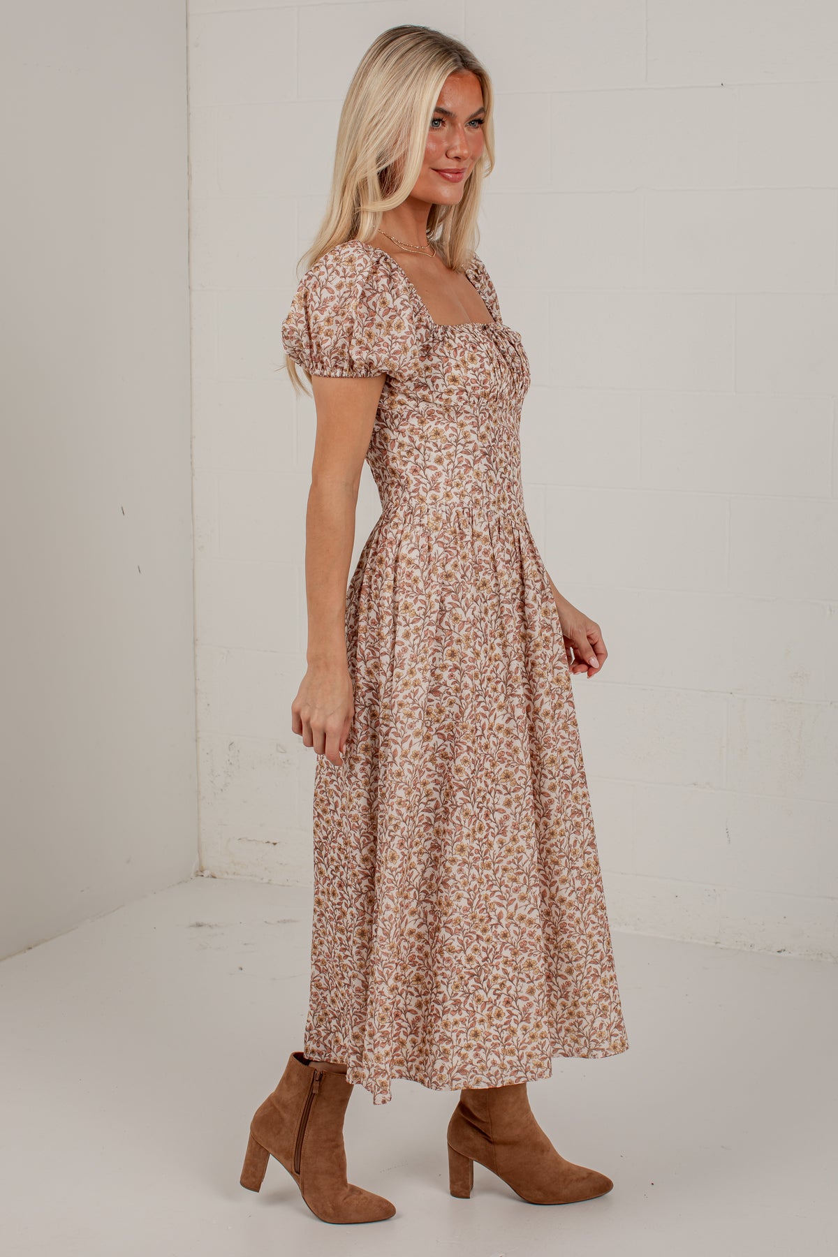 A woman modeling a midi-length, puff-sleeve dress with a small floral print in beige and brown tones, paired with brown suede ankle boots, standing against a white brick wall.