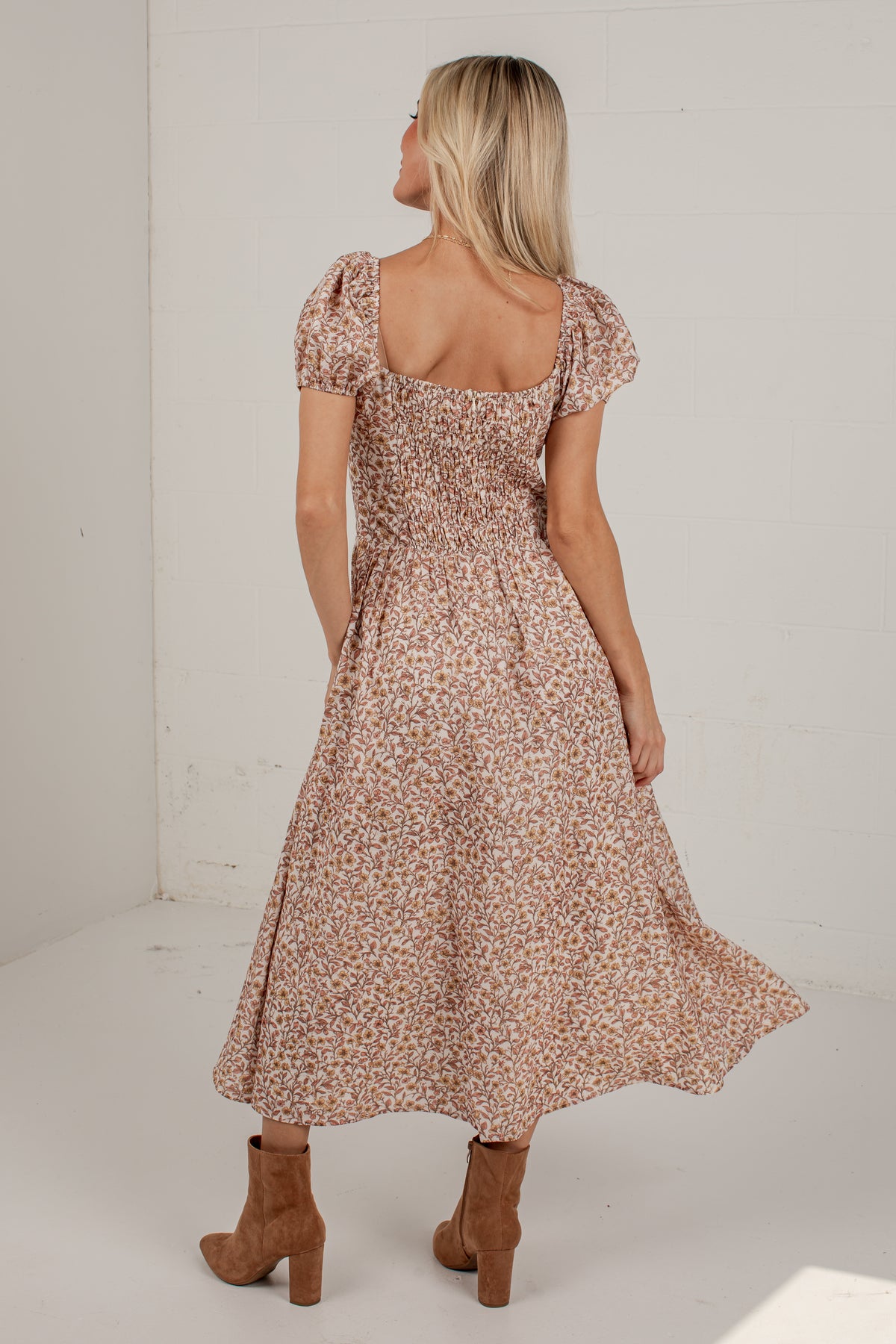 A woman wearing a brown and beige floral print midi dress with puff sleeves and a smocked bodice, shown from the back in a bright studio setting.
