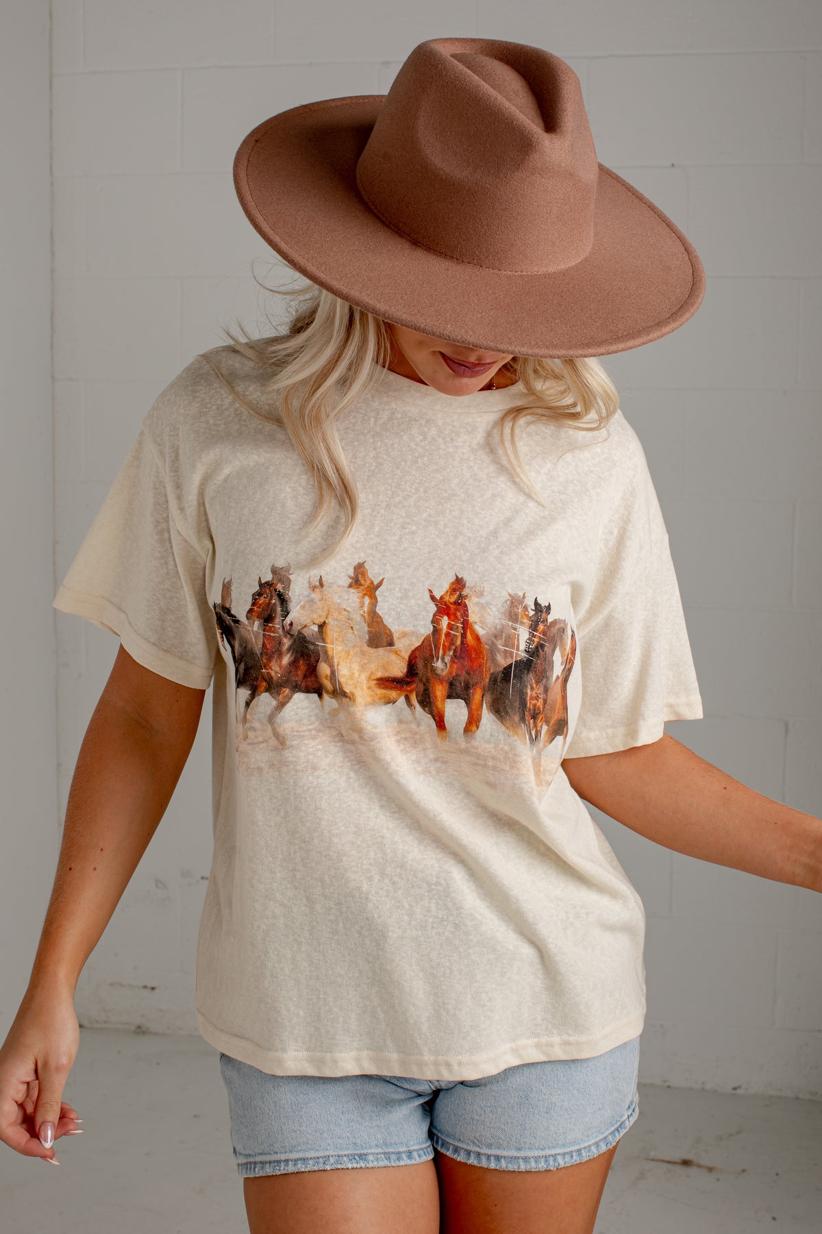 A woman wearing a cream-colored short-sleeve t-shirt with a western-themed horse and rider graphic, paired with denim shorts and a brown wide-brim hat, standing against a white brick wall.