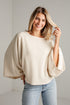 A woman wearing a cream-colored knit poncho sweater with wide flared sleeves and light blue jeans, standing against a white paneled wall.