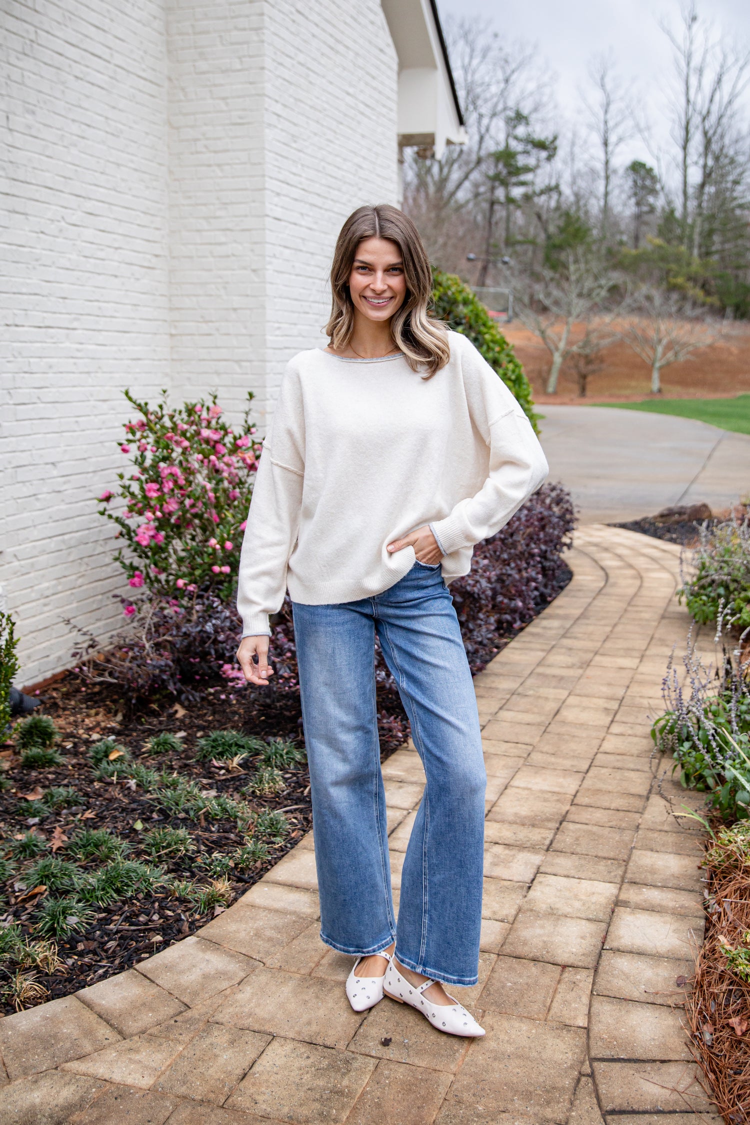 Woman in a white sweater and blue jeans standing on a brick path with plants and flowers in the background.
