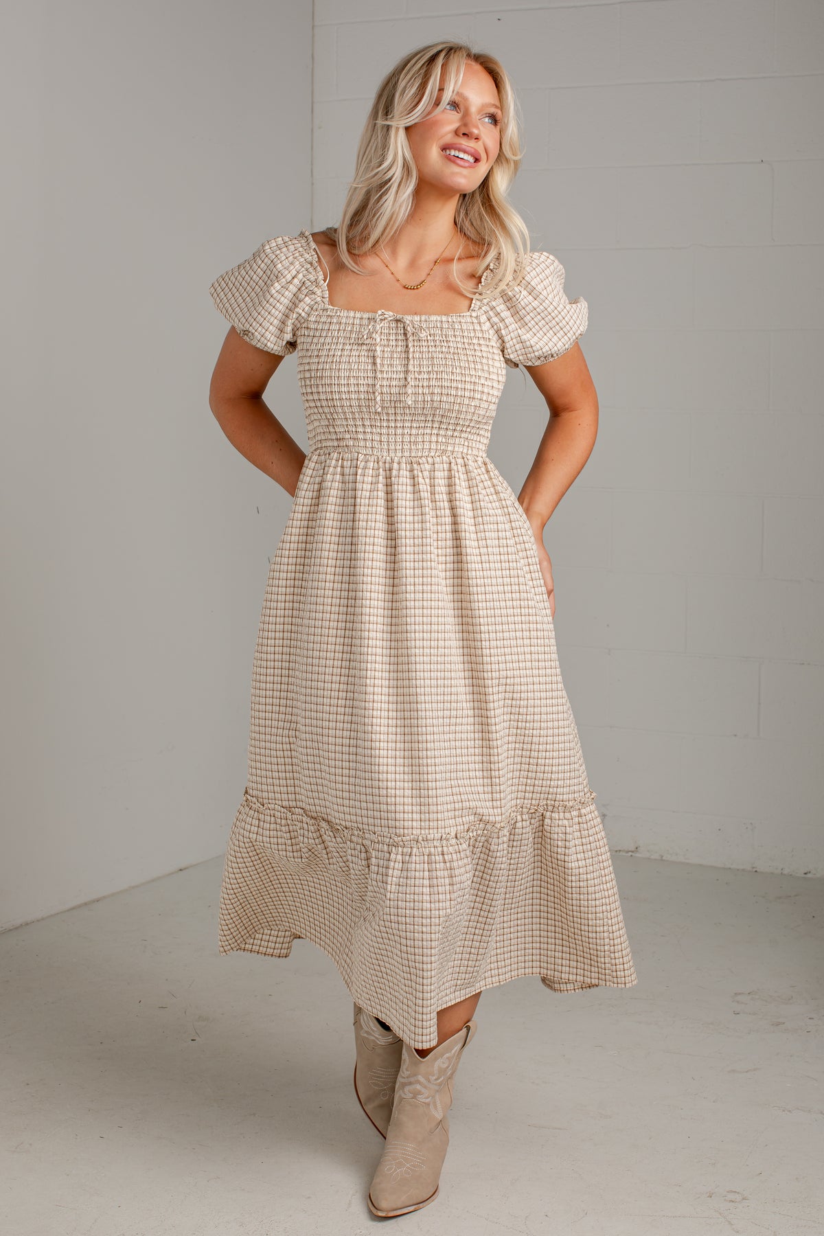 A woman modeling a beige and white gingham tiered midi dress with puff sleeves and a smocked bodice, paired with beige ankle boots, in a minimalist studio setting.