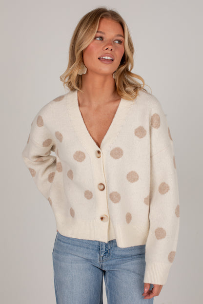 A woman wearing a cream-colored knit cardigan with beige polka dots and light blue jeans, photographed against a neutral light gray studio background.