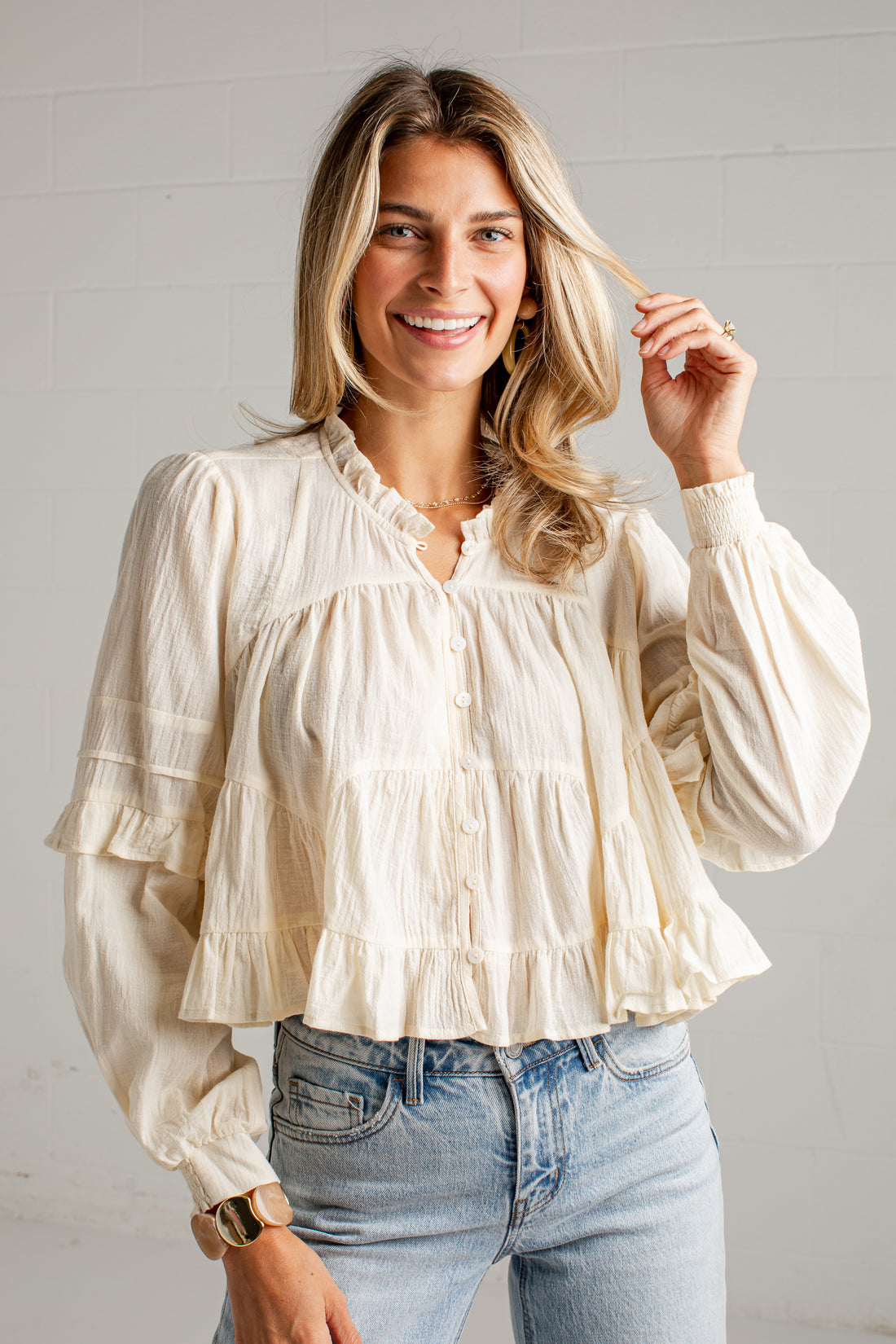 A woman wearing a cream-colored, long-sleeve tiered blouse with ruffle details and light-wash blue jeans, standing against a white brick wall.
