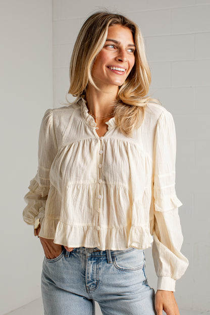 A woman wearing a cream-colored, long-sleeve crinkle blouse with a ruffled collar and button front, paired with light-wash blue jeans, standing against a white brick wall.