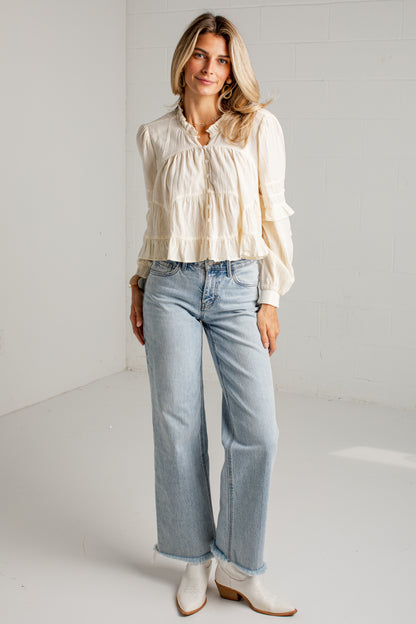 A woman stands in a bright white studio wearing a cream ruffled long-sleeve blouse, light-wash wide-leg jeans, and white ankle boots.