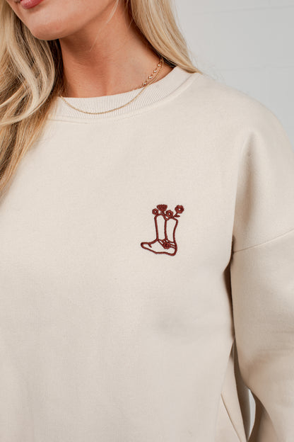 A close-up of a woman wearing a cream-colored crewneck sweatshirt with a small embroidered graphic of a sock on the left chest.