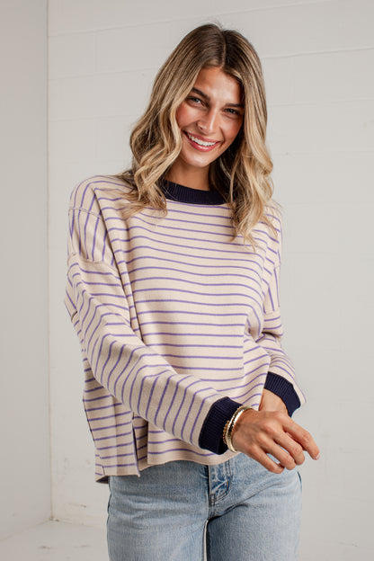 A woman wearing a lavender and navy blue horizontally striped long-sleeve knit sweater with solid navy ribbed cuffs and collar, paired with light-wash blue jeans, standing against a white paneled wall.