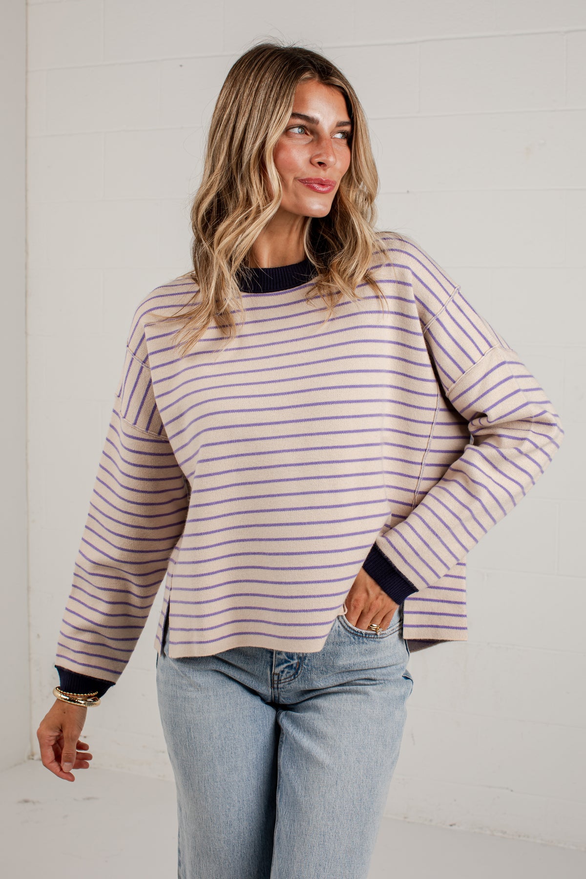 A woman wearing a long-sleeve, off-white ribbed knit sweater with thin purple and navy horizontal stripes, paired with light-wash blue jeans, standing against a white paneled wall.
