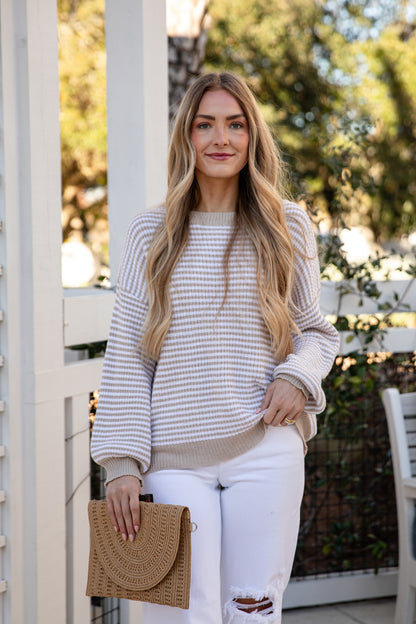 Woman standing outdoors holding a beige handbag, wearing a striped sweater and white pants.