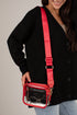 A woman wearing a black cardigan and light blue jeans carries a small transparent waterproof pouch bag with a bright red strap and red trim. The pouch contains round sunglasses.