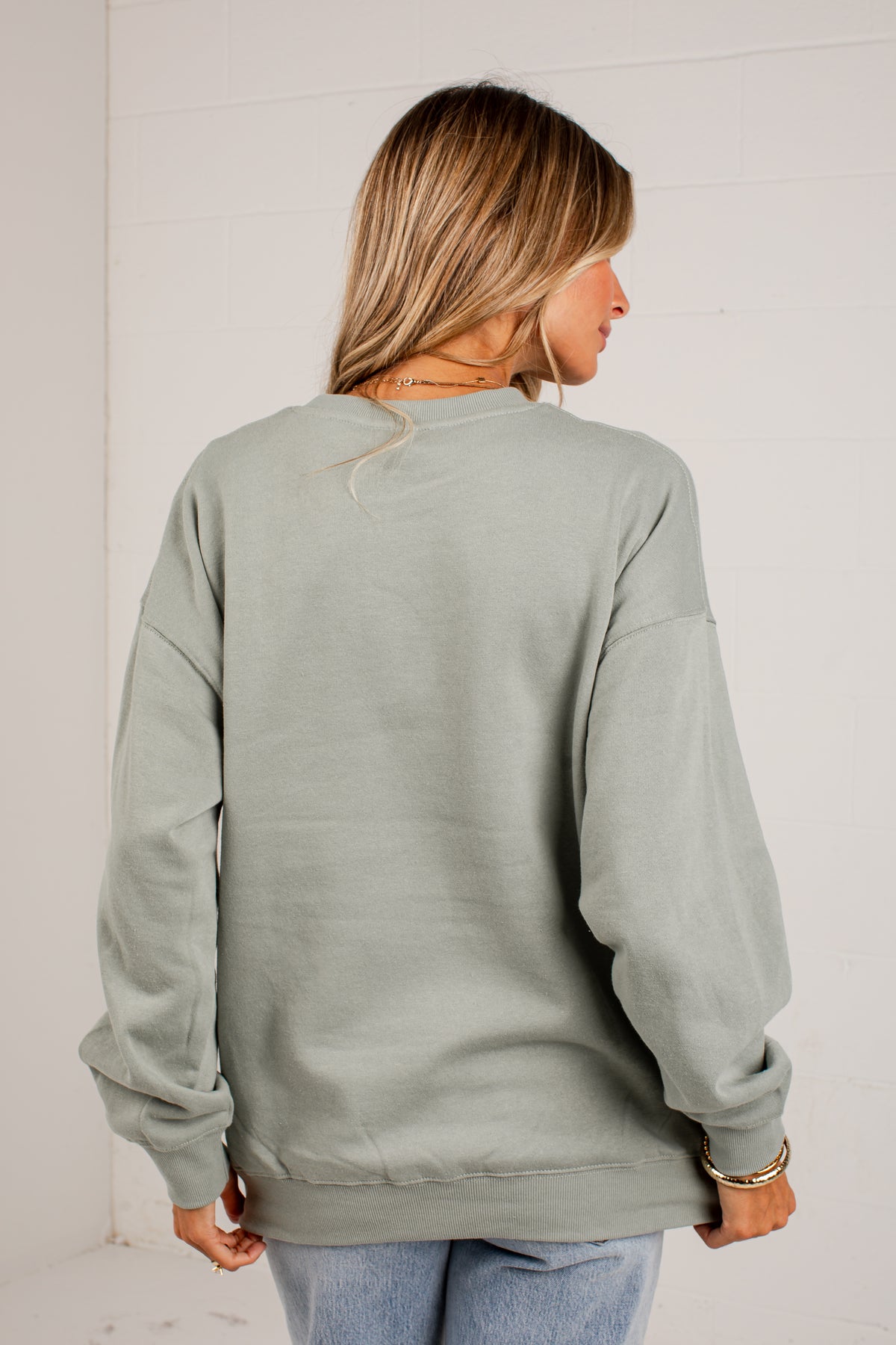 A woman viewed from the back wearing an oversized sage green crewneck sweatshirt and light blue jeans, standing against a white paneled wall.